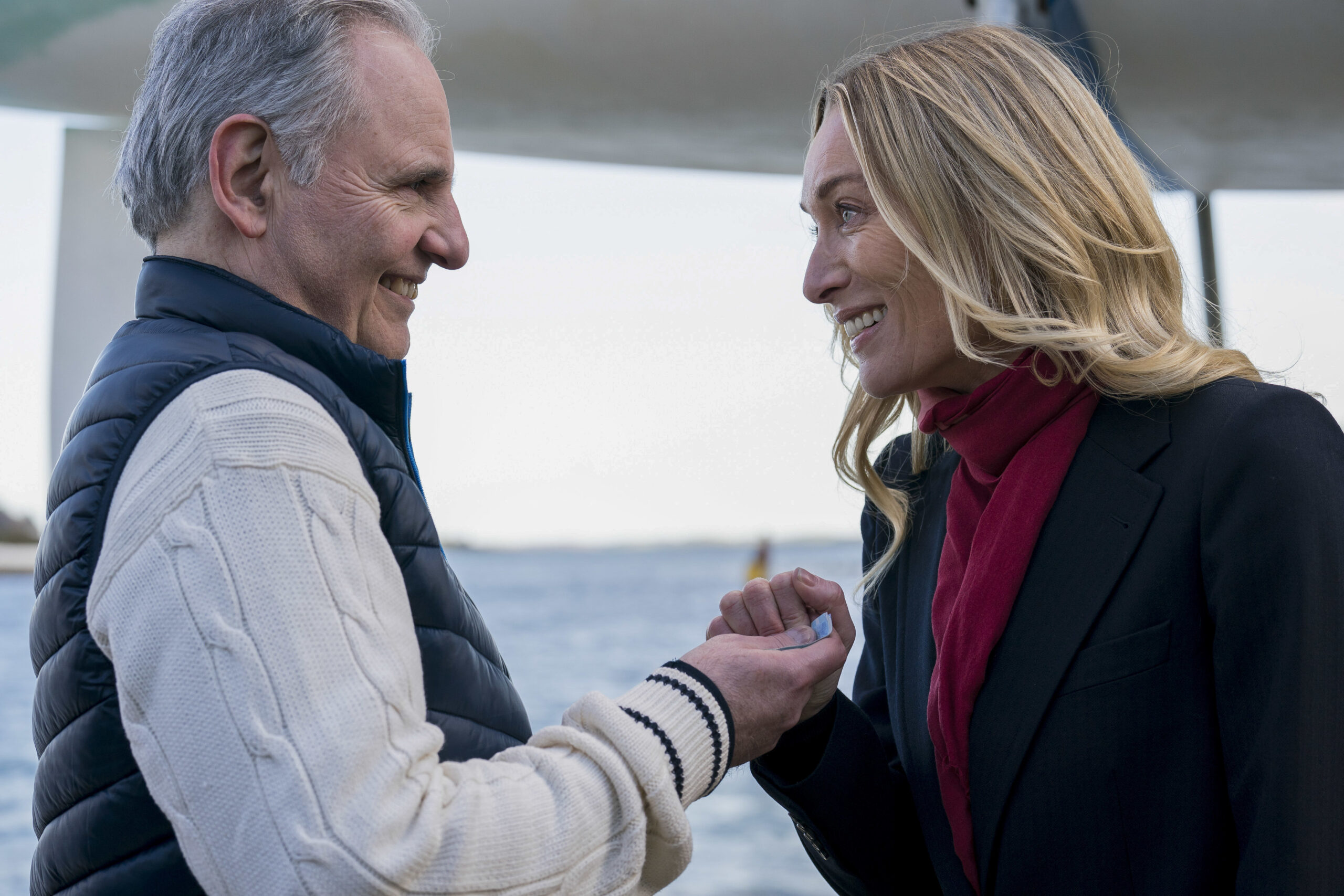 Richard Coxford as Yacht Club Sec and Victoria Smurfit as Olivia Foyle, they face each other both smiling next to a boat