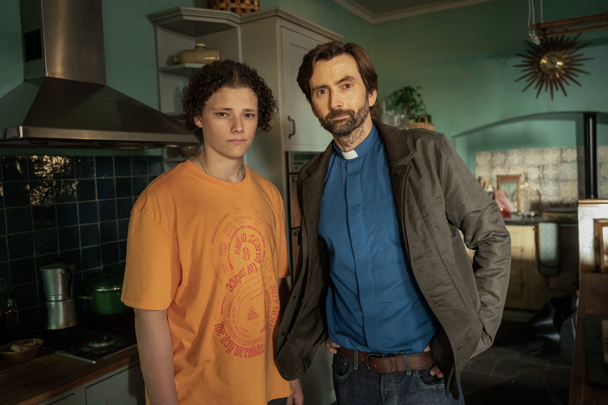 Louis Oliver as Ben and David Tennant as Henry Watling, they stand in the watling family home with frowns on their faces 