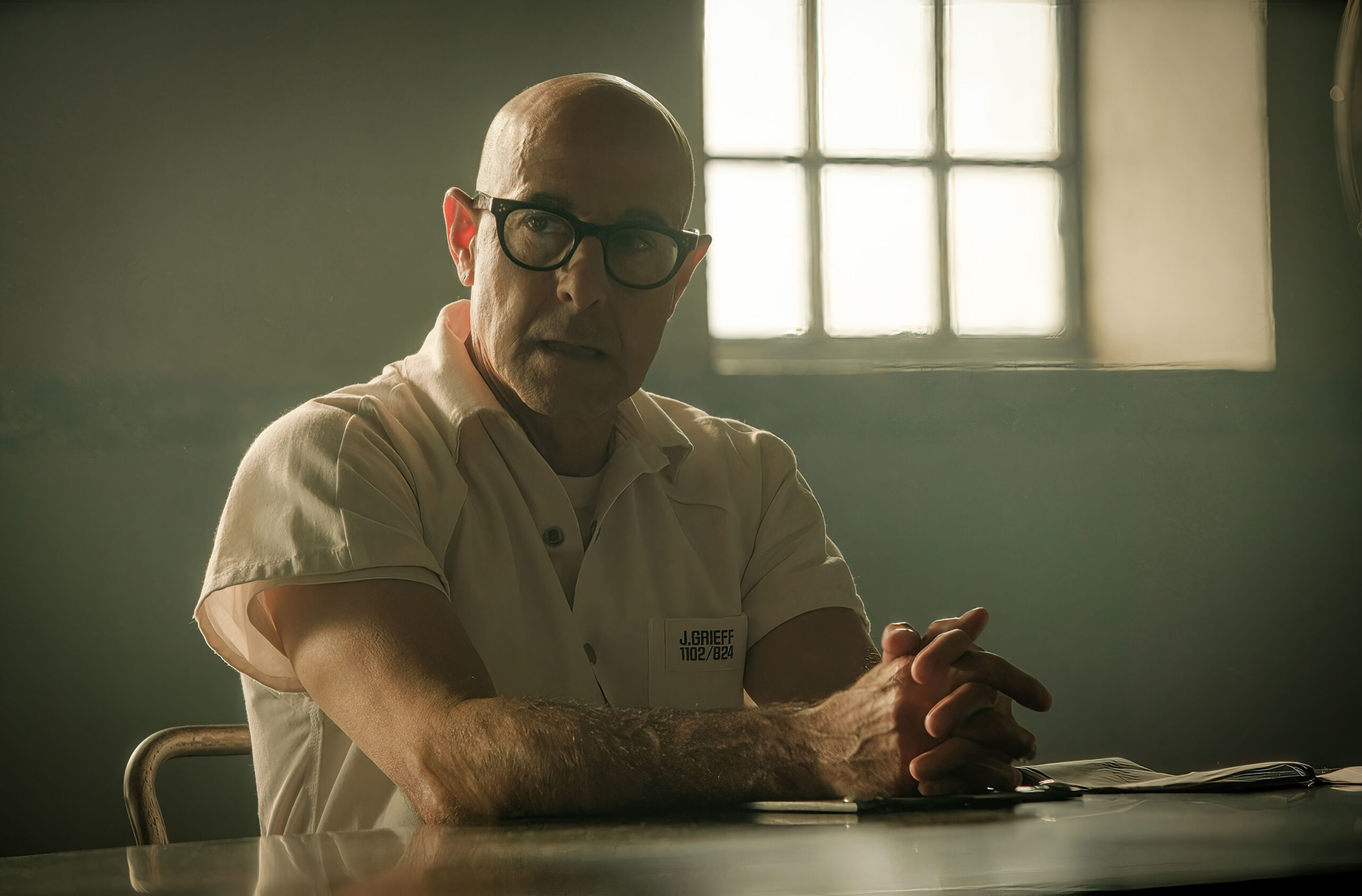 Stanley Tucci as Jefferson Grieff in Inside Man, he sits at a prison table in a prison cell with a shocked expressions