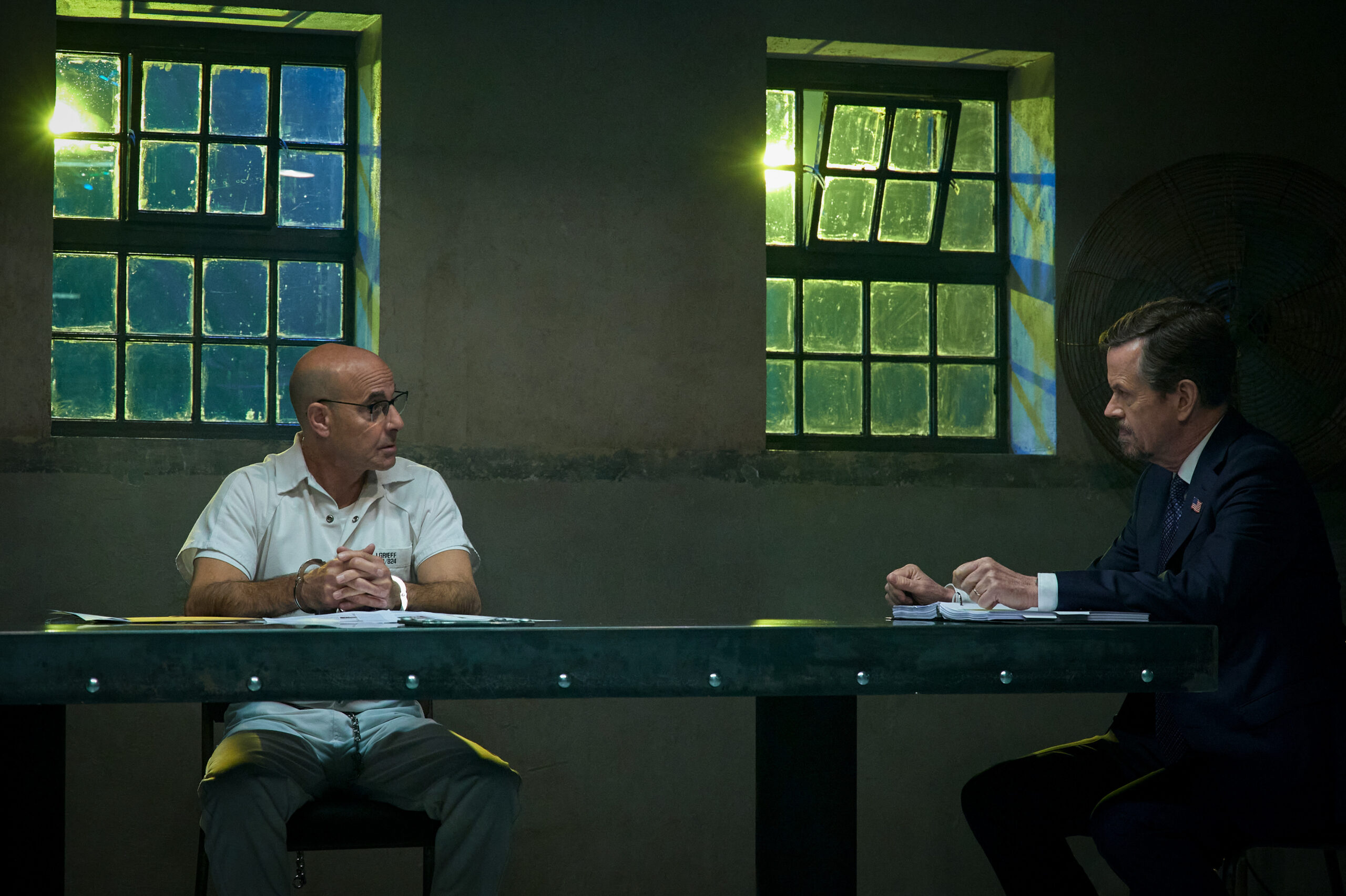 Stanley Tucci as Jefferson Grieff (left) and dylan baker as warden casey (right) they sit at a table in the prison with concerned expressions