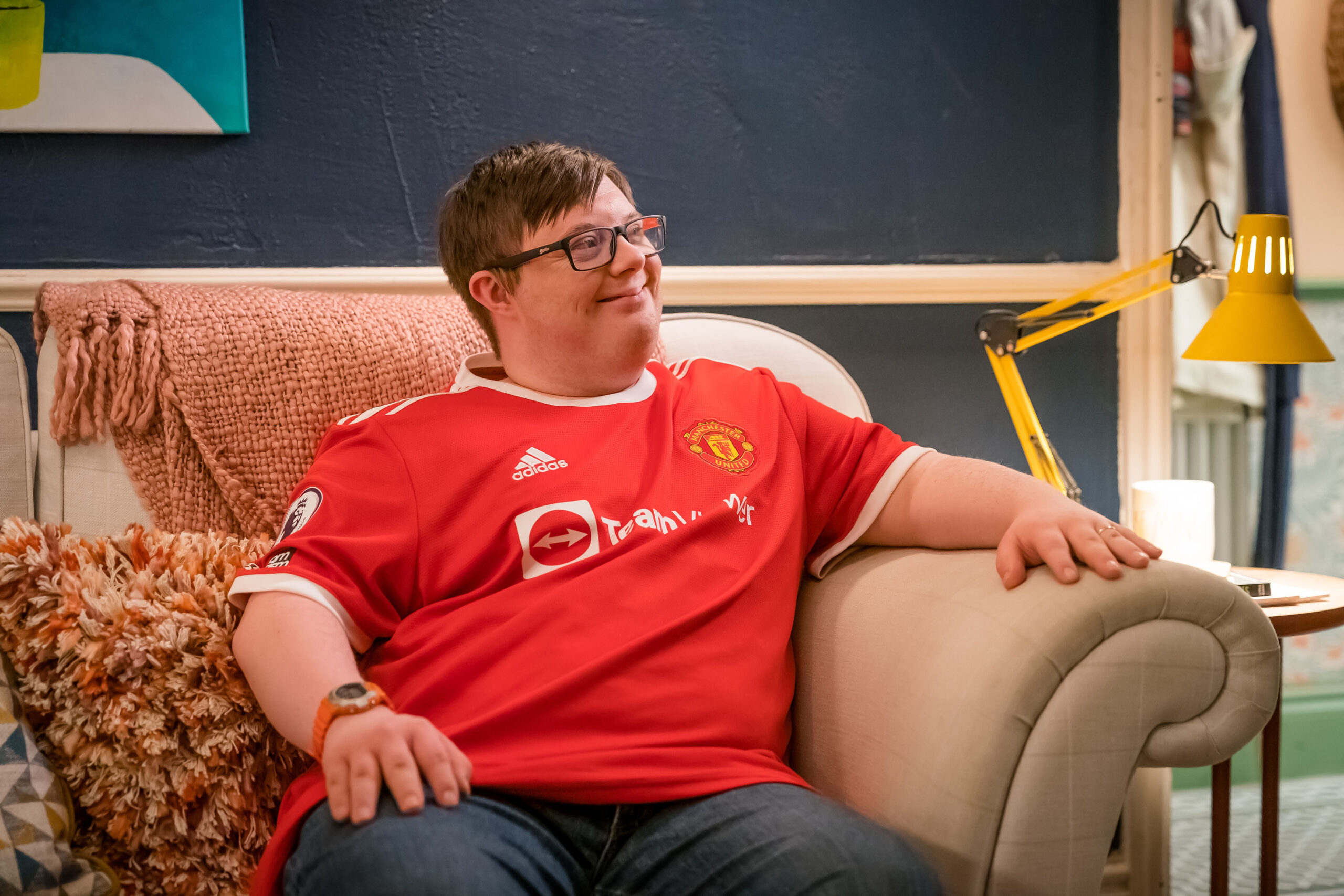 Leon Harrop as Ralph Wilson in Ralph & Katie, he sits on a sofa with a smile and a Man UTD top on