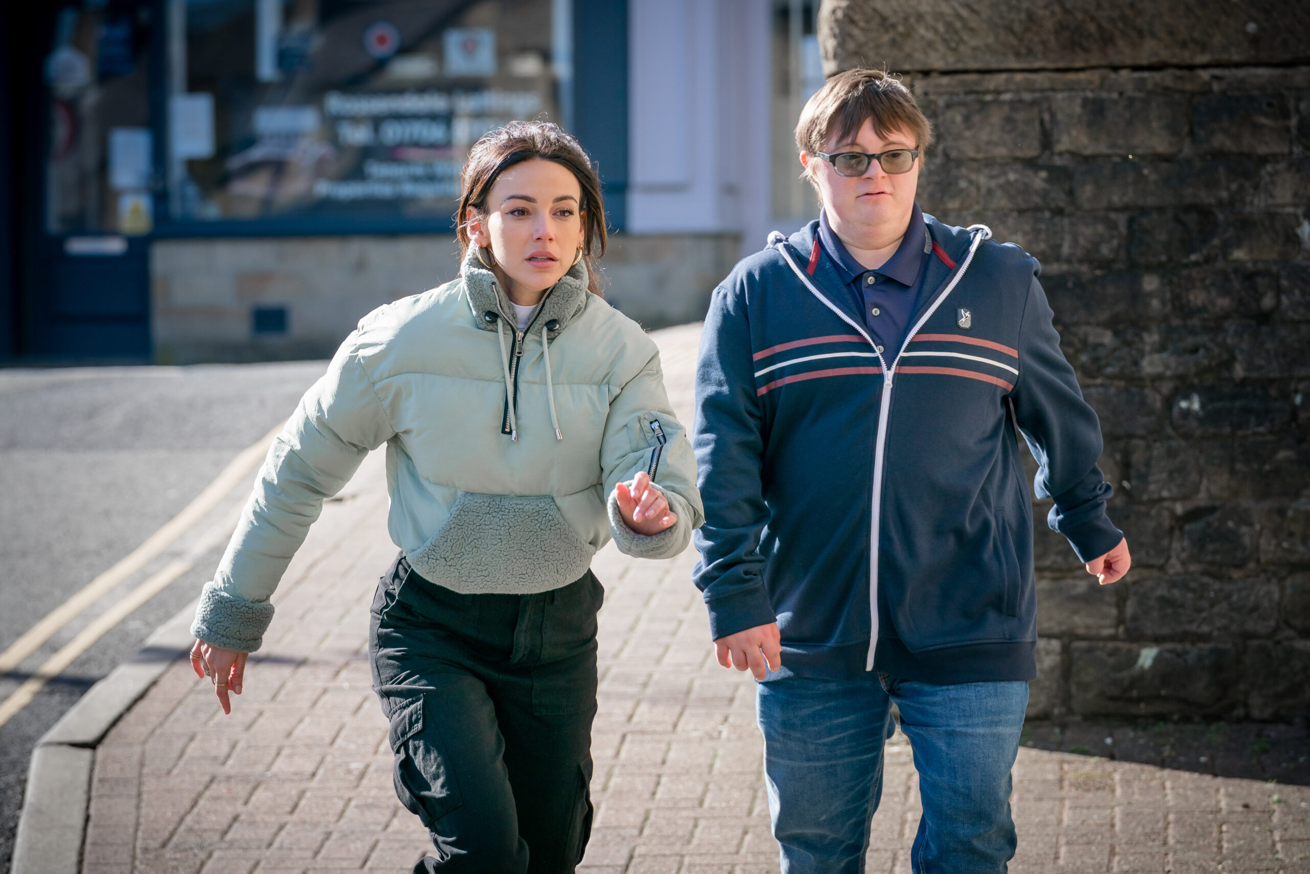 Michelle Keegan as Erin and Leon Harrop as Albie in Brassic, they walk down a street with stressed faces