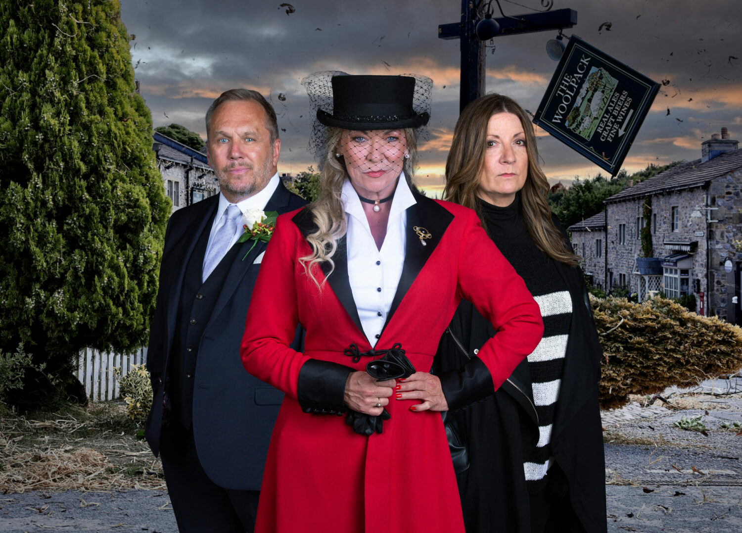 Emmerdale promo picture, Will in wedding suit, Kim in a red cloak and top hat and Harriet in a stripy jumper, all looking serious