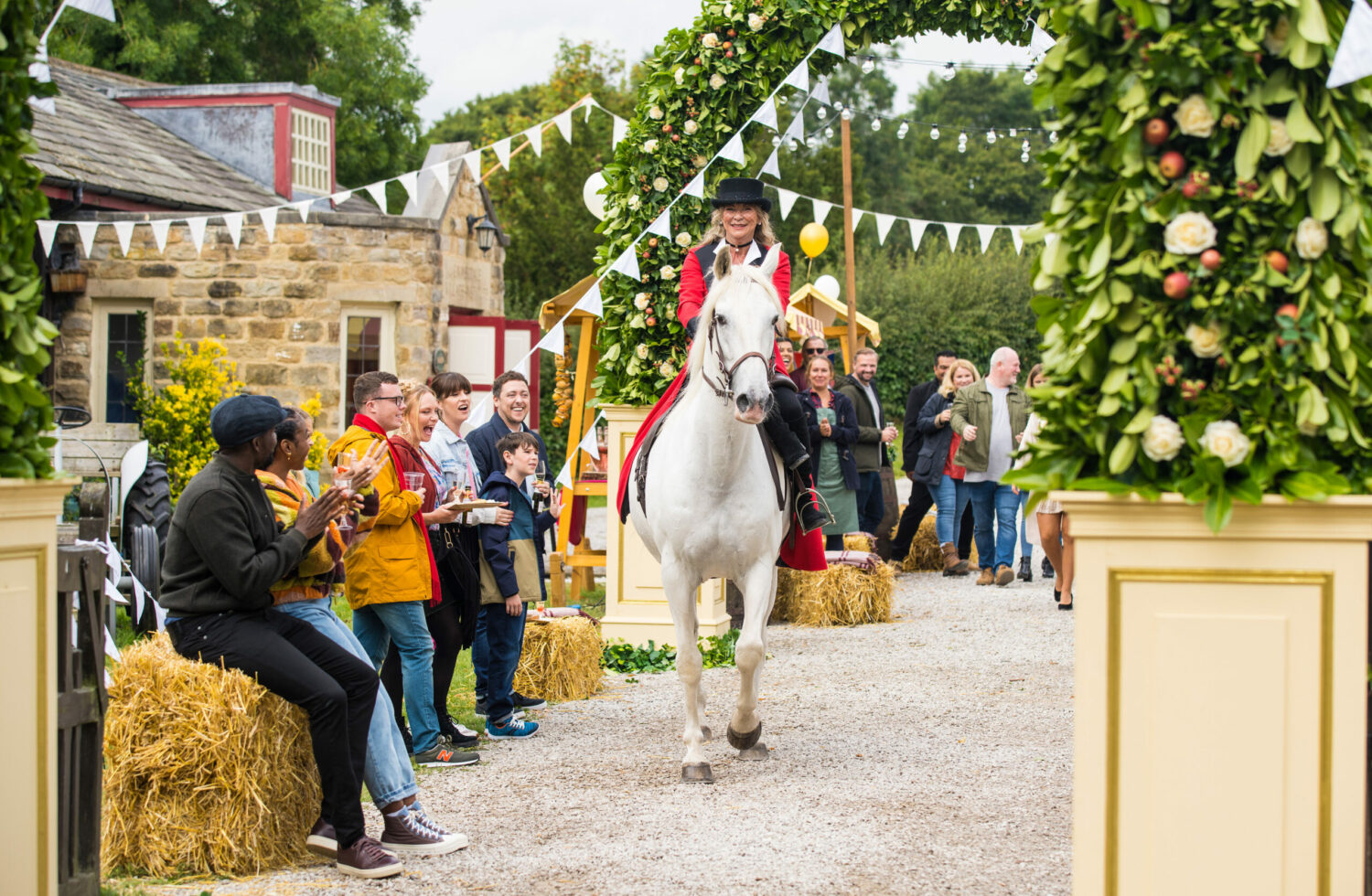 Emmerdale villagers celebrating as Kim wears a red cloak and rides a white horse through the village