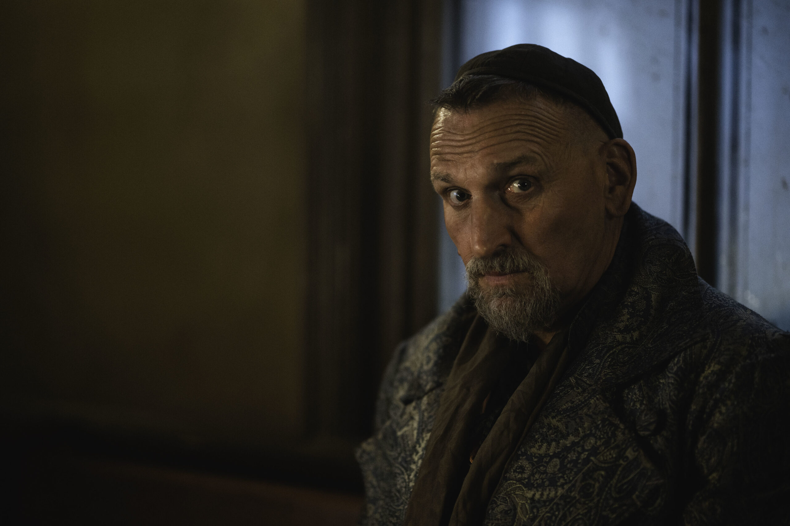 Christopher Eccleston as Fagin in Dodger, he looks into the camera with a worried expression he wears rough period clothing in a dark room