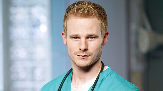 Steven Miller as Lenny Lyons in Casualty. He smiles in scrubs standing next to a white wall