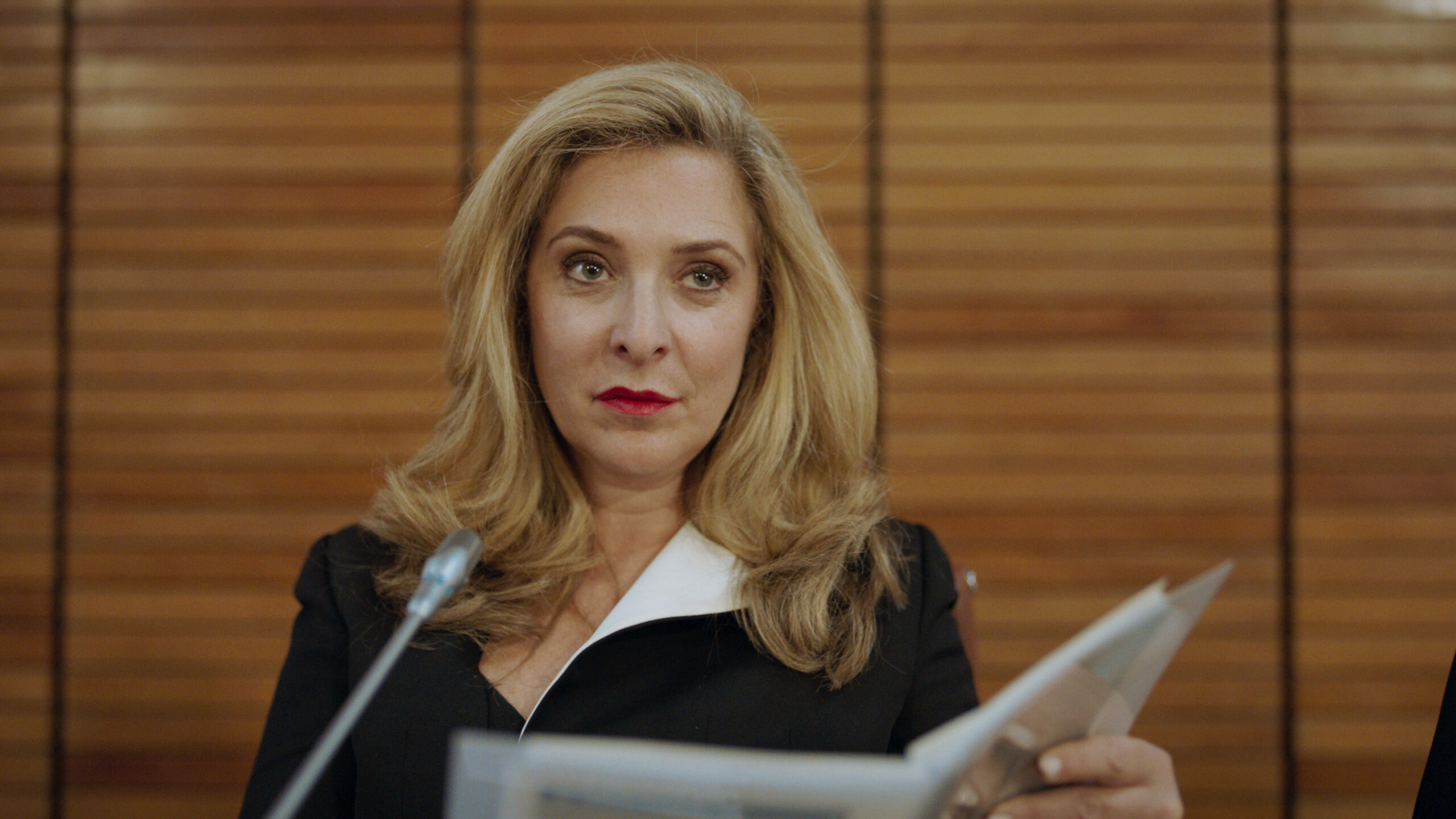 Tracy-Ann Oberman as Helen Chalmers in Code 404, she smiles as she reads from a booklet next to a mic, she is sitting down 