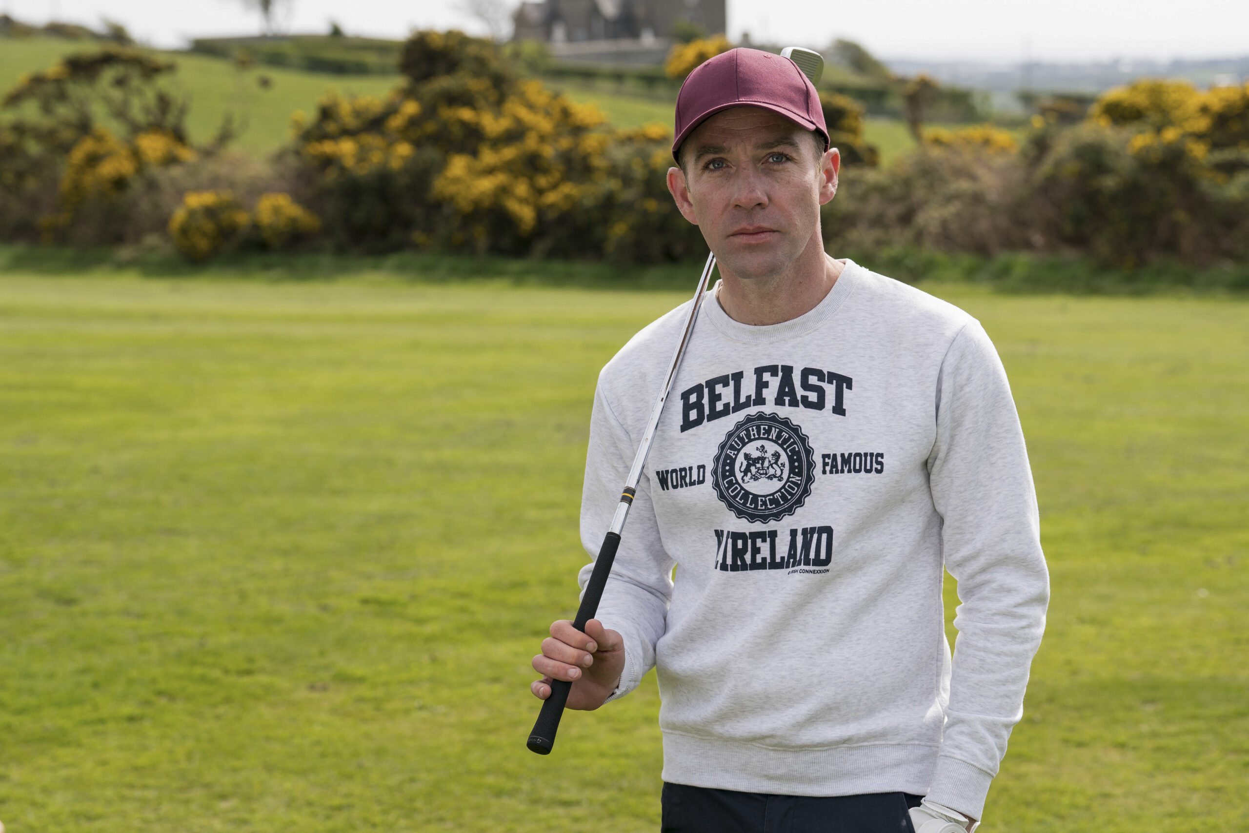 Jonjo O'Neill as Ryan Savage in Bloodlands, he holds a gold club and looks into the camera with a small smile with lots of grass and flowers behind him