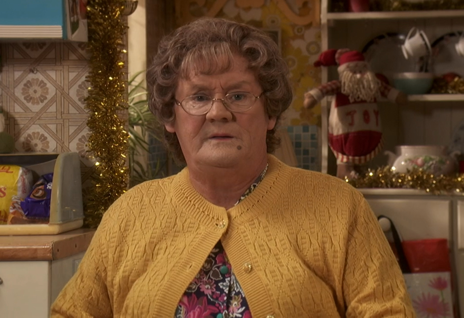 Brendan O'Carroll as Mrs Brown in Mrs Brown's Boys, she has a grumpy look as she sits in a kitchen in a yellow cardigan