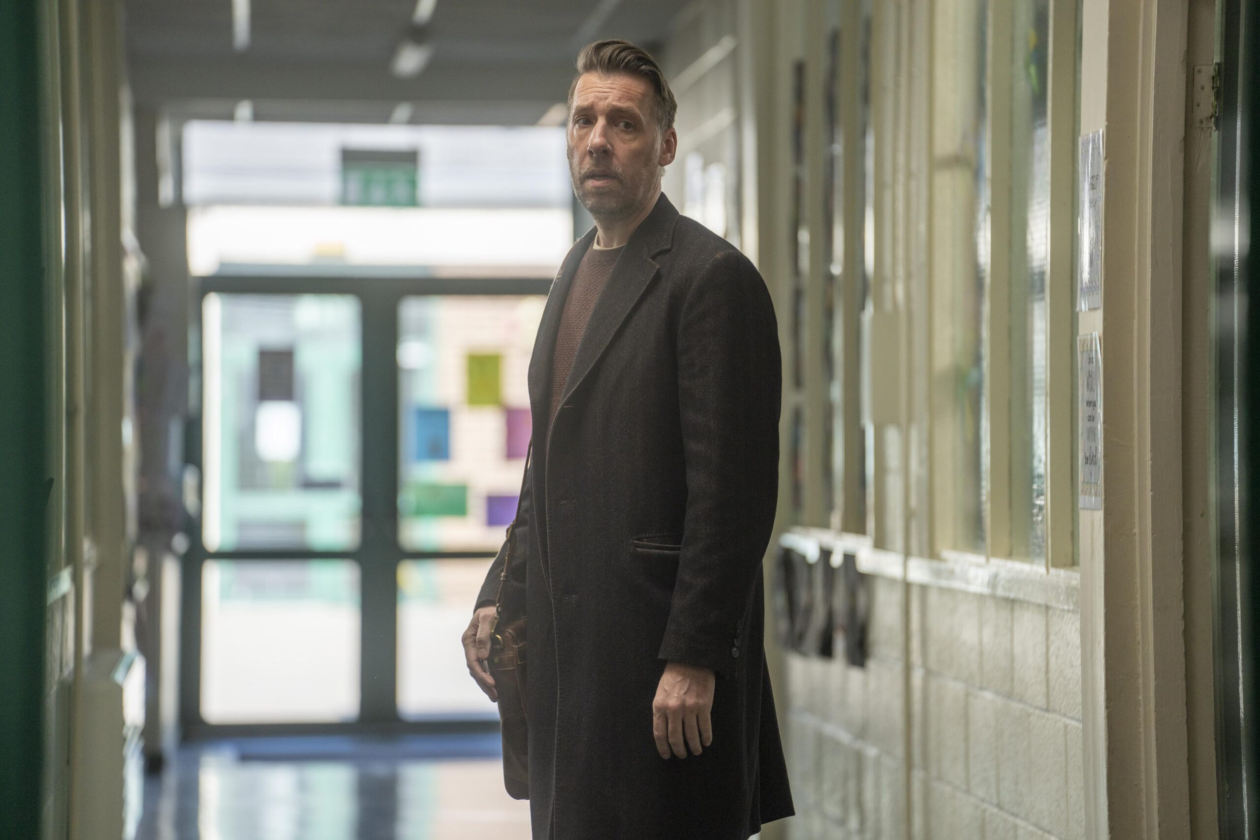 Craig Parkinson as George in The House Across the street, he looks down a long school corridor with a frown