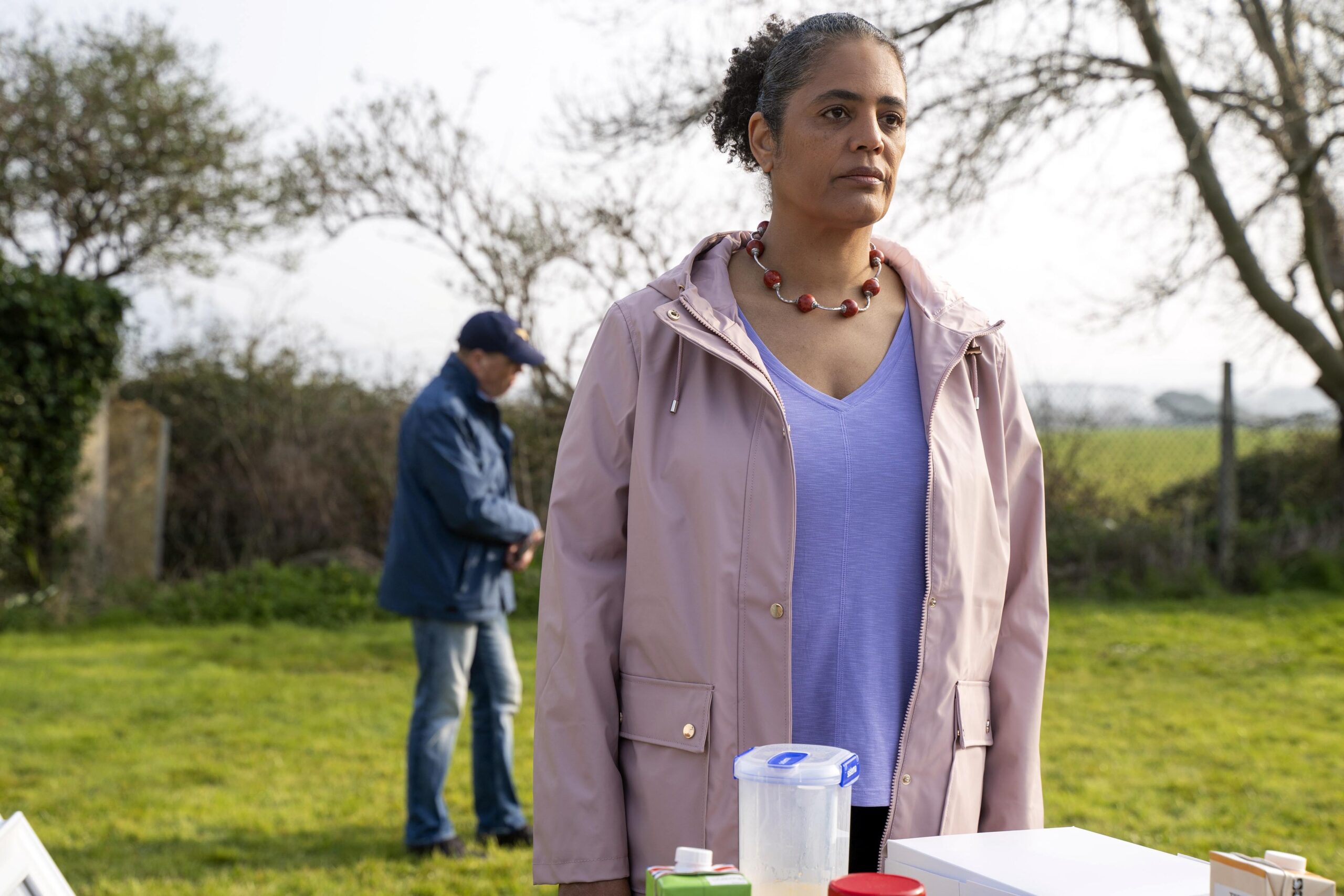 Sara Powell as Joanne, she stands outside in a pink coat with a worried expression