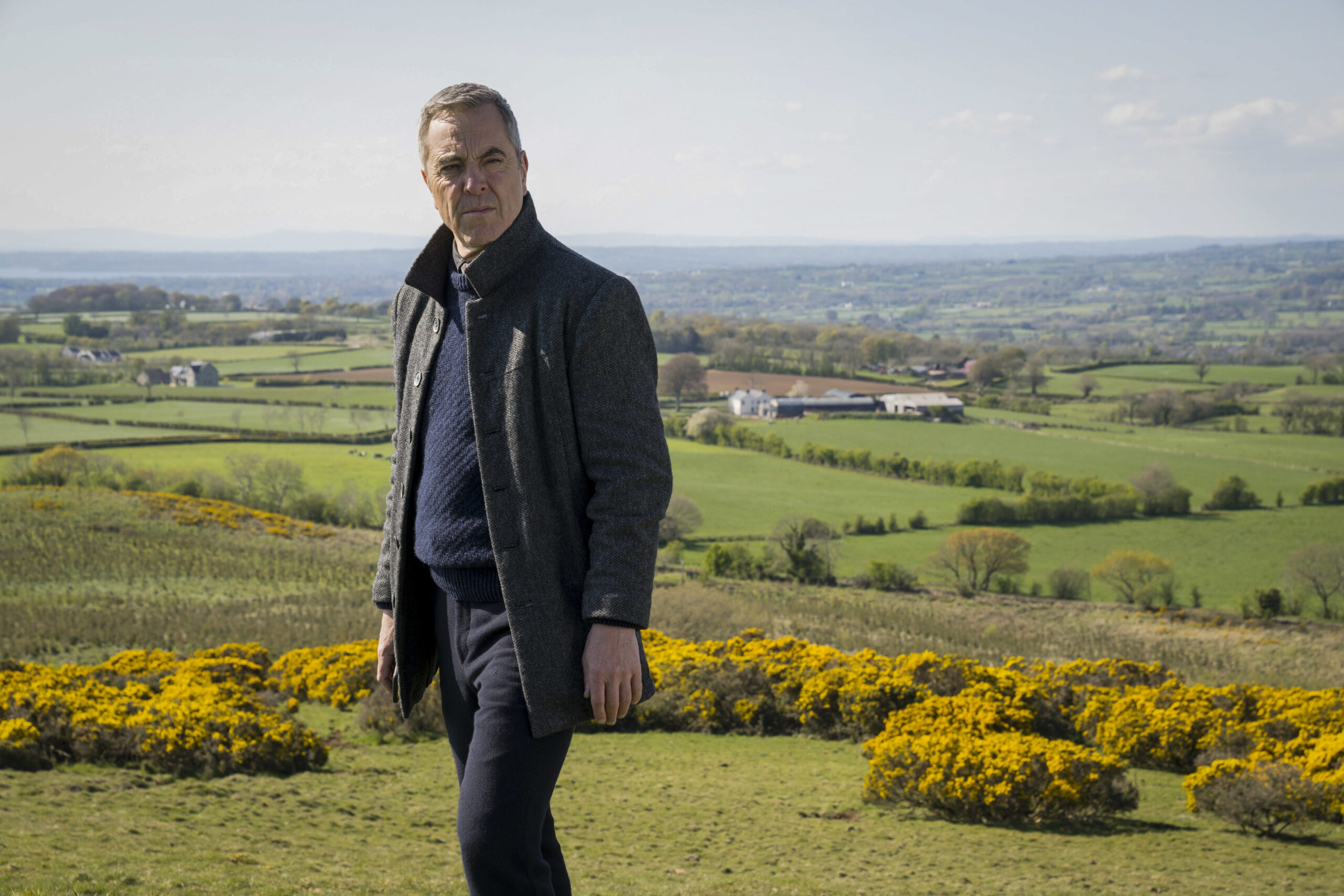 James Nesbitt as Tom Brannick in Bloodlands, he looks to his right with a concerned facial expression he stands on a big green hill