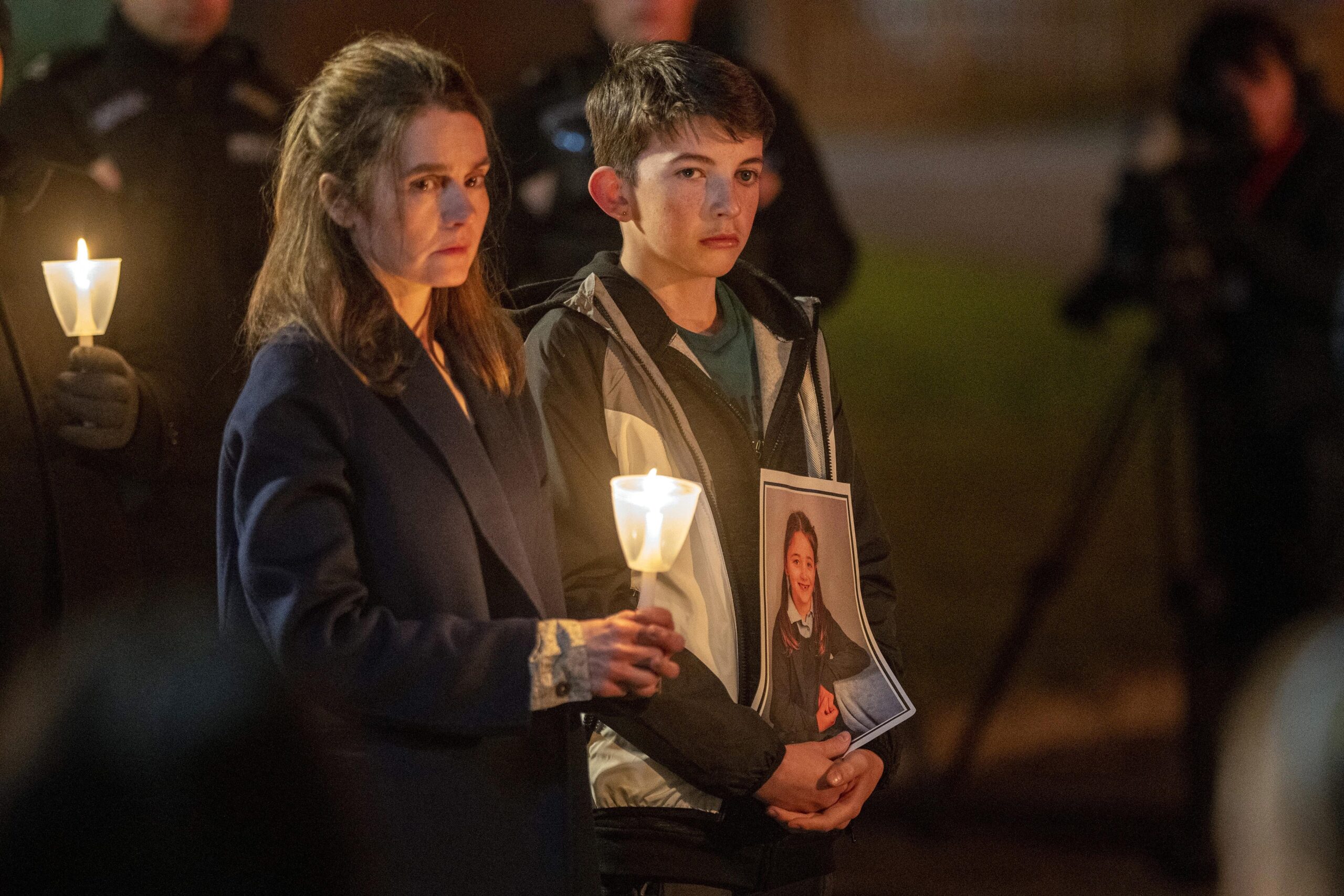 Shirley Henderson as Claudia and Caleb Wilson as Rhys, they stand at the vigil with sad faces and candles in their hands