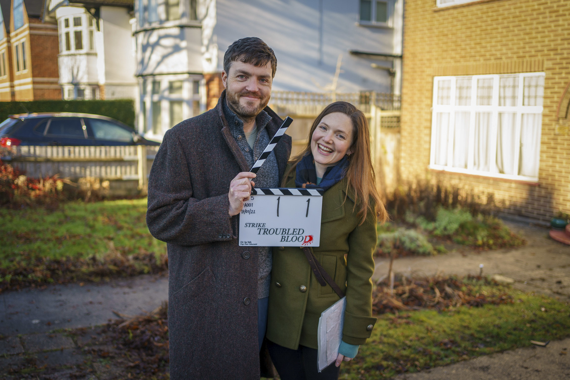 Tom Burke and Holliday Grainger smiling happily as they hold filming clapper for Strike