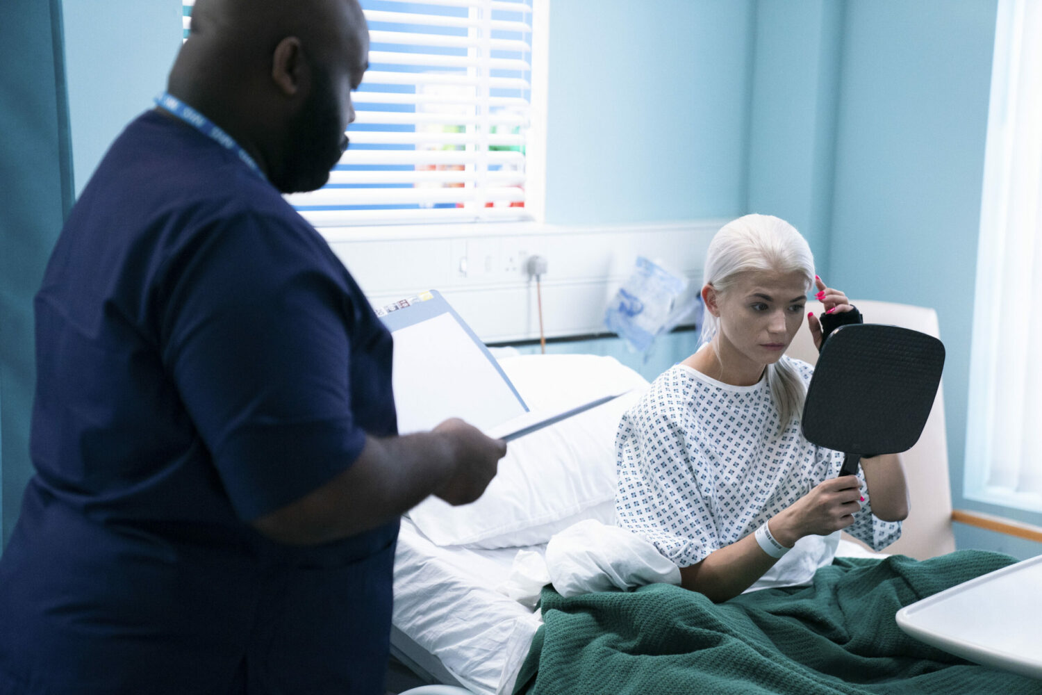 EastEnders Lola holding a mirror in a hospital bed while a consultant talks to her