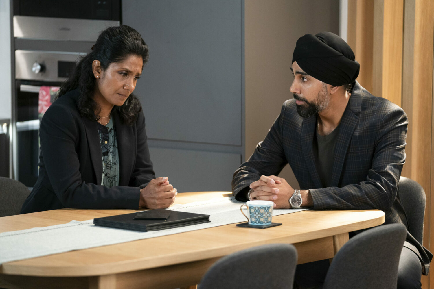 Suki and Kheerat in EastEnders chat solemnly at a table 