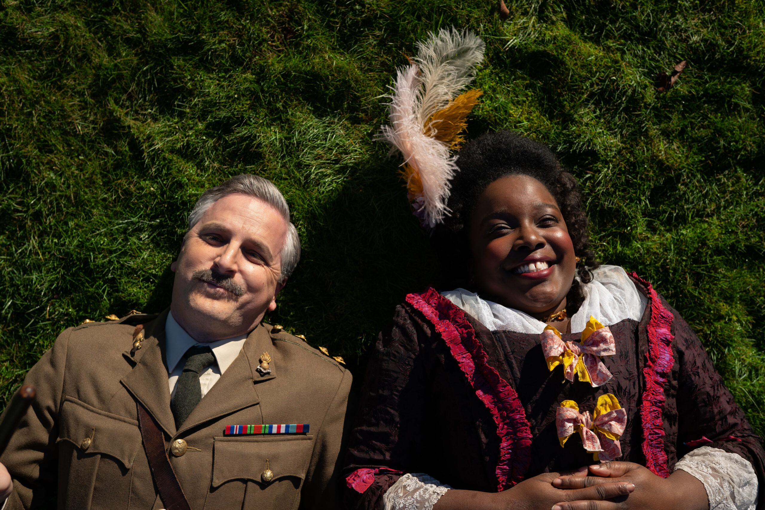 Captain (BEN WILLBOND), Kitty (LOLLY ADEFOPE) in Ghosts, they look up at the sky with smiles as they lay on grass