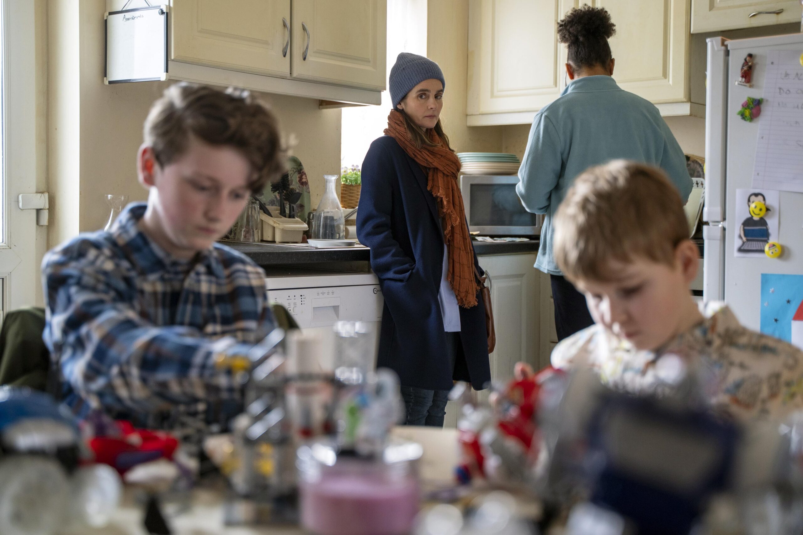 Calum Jess as Dean and Eddie Brett as Simon, they both have their heads down as they sit at the table with frowns