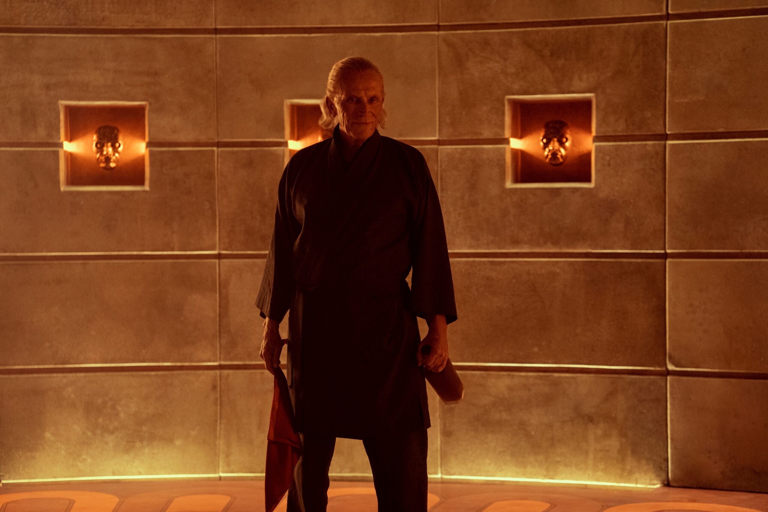 Peter Weller stands in front of a golden wall, holding a bottle of red liquid and dressed in a black robe