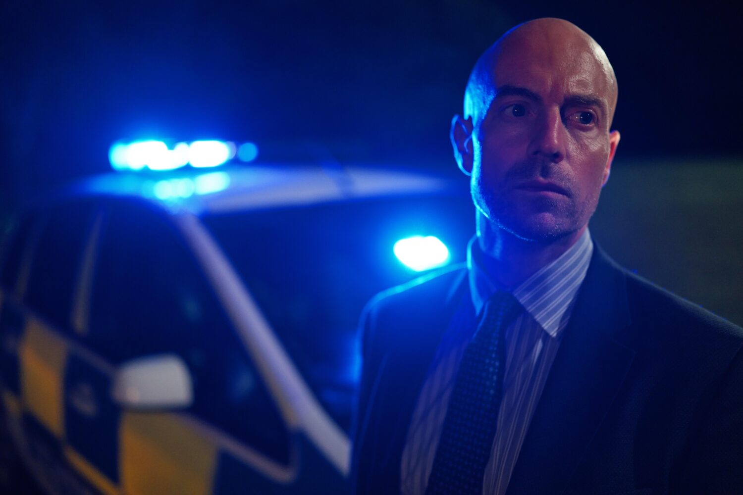 Matthew Gravelle as DS Pritchard in The Pact series 2 standing in front a a blue light on police car looking worried