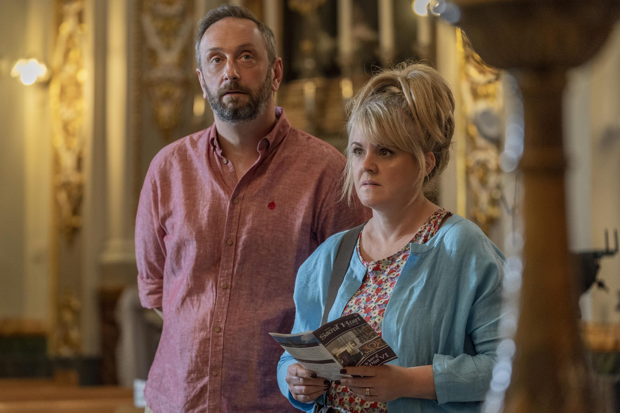 (LEFT) Steve Edge as Dom and (RIGHT) Sally Lindsay as Jean White in The Madame Blanc Mysteries - they stand in a church with confused expressions