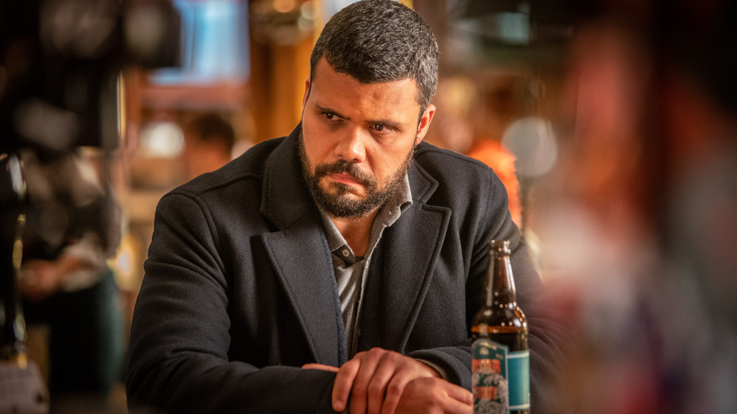 Howard Charles as Mike McGuire in Whitstable Pearl, he sits at a table for a frown with a beer bottle on the table next to him