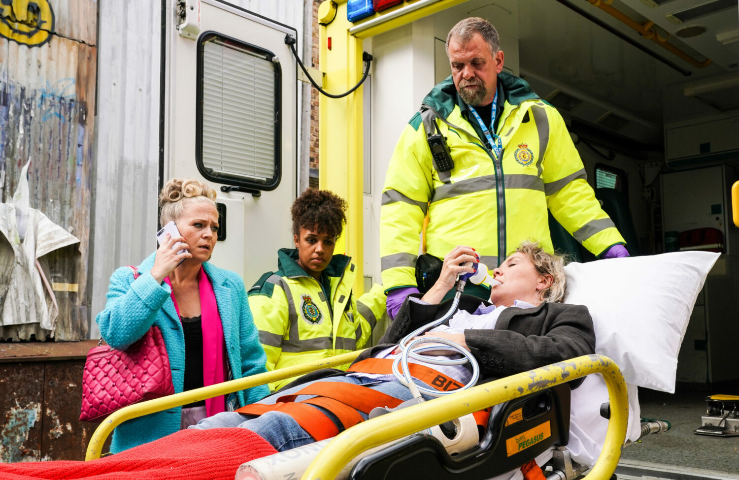 Linda looks worried on the phone as Janine lies outside of an ambulance with paramedics around her in EastEnders