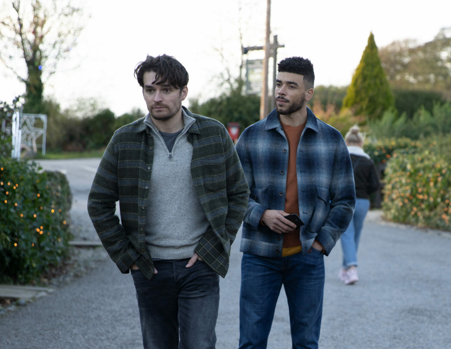 Nate watches Mack suspiciously as they walk in the street on Emmerdale