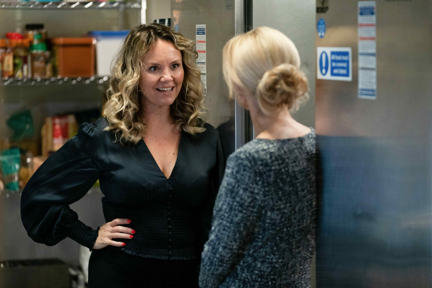 Janine Butcher smiles with her hand on her hip as she talks to Kathy Beale in the kitchen