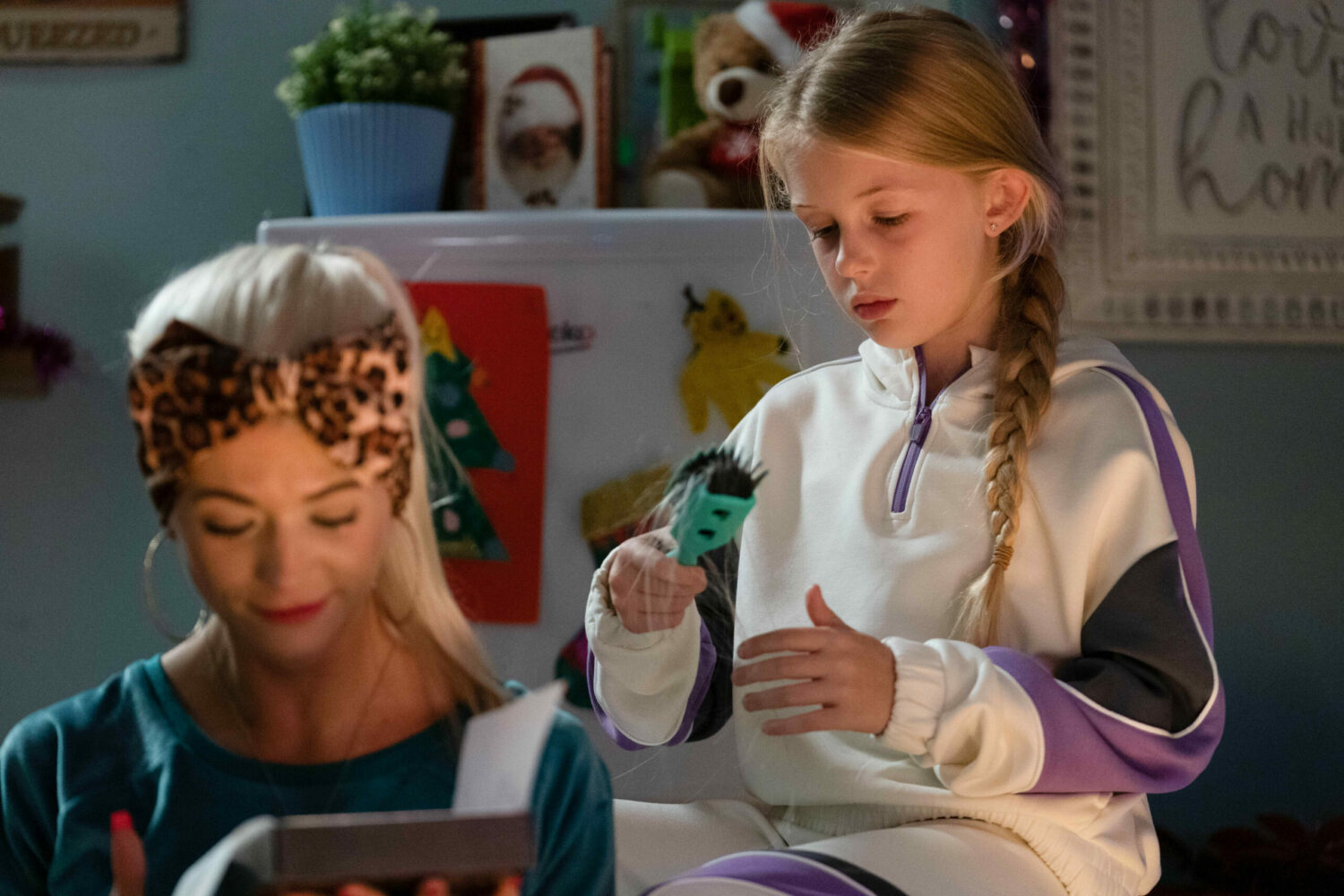 Lexi looking at a hairbrush as she sits next to a smiling Lola in EastEnders