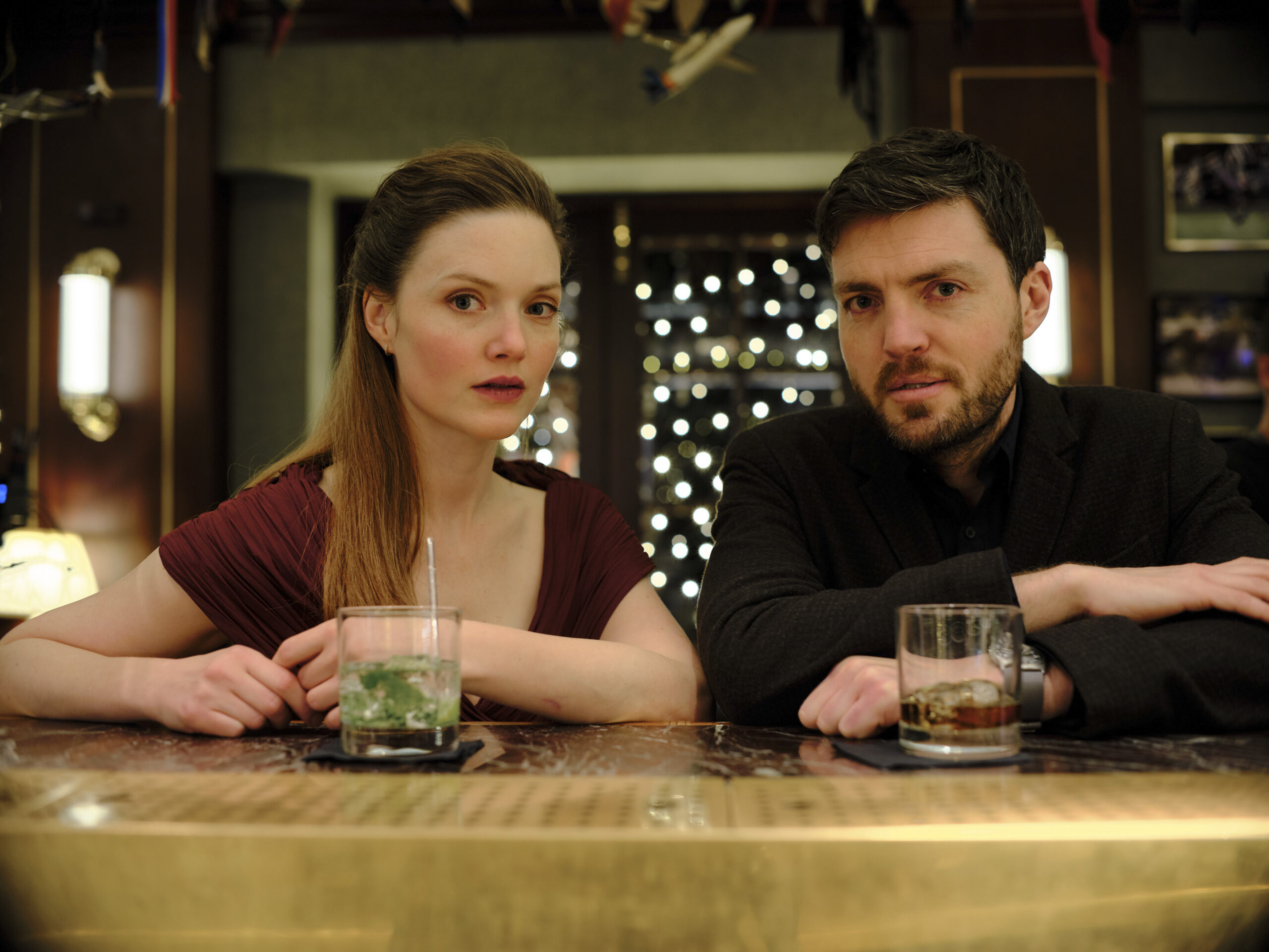 Robin and Strike sit close at a bar