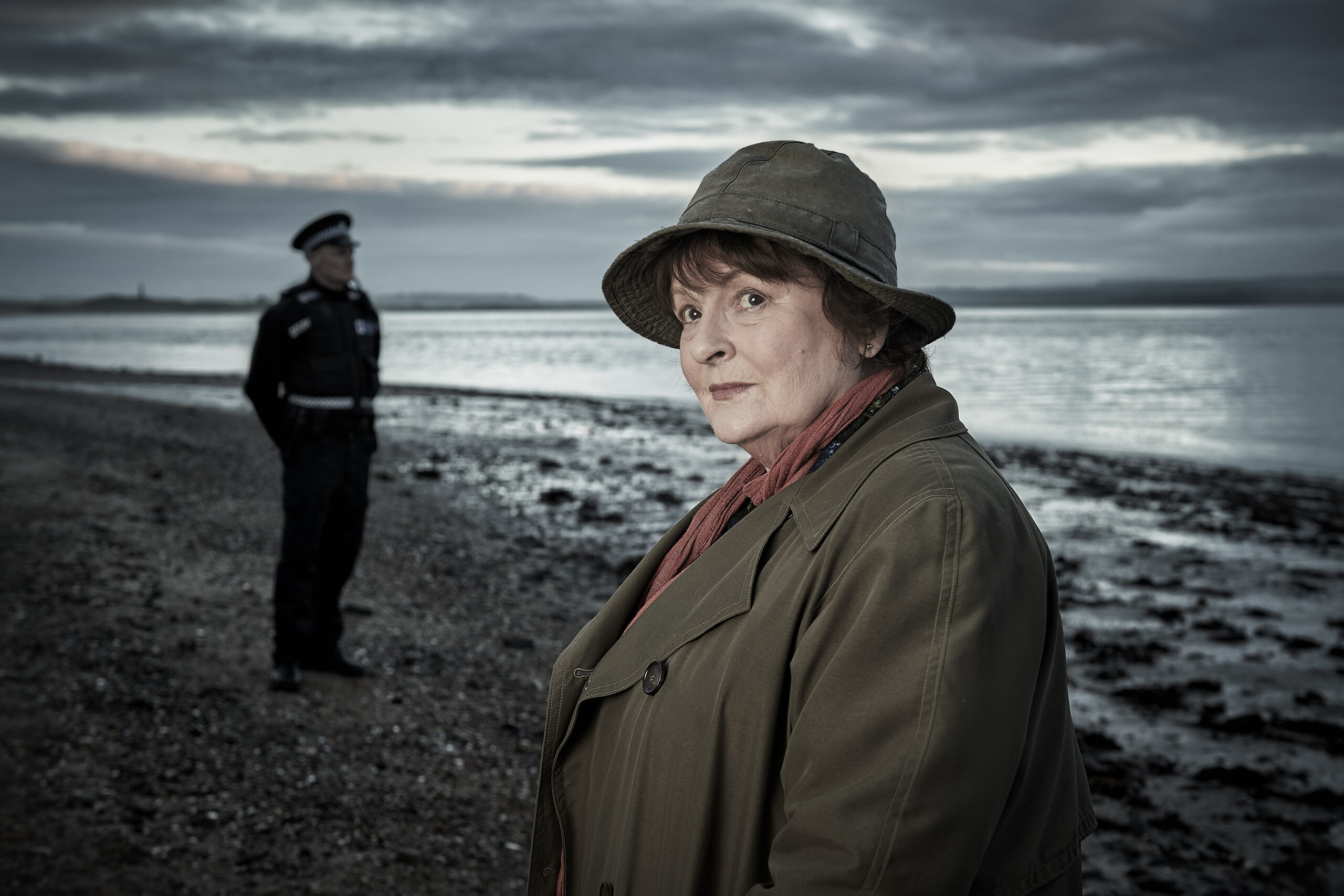 Vera stands on a beach with a police officer behind her in series 12