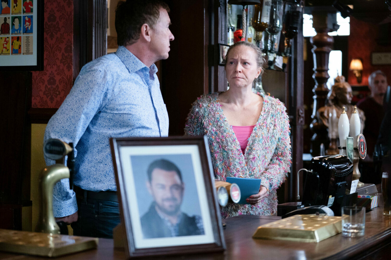 Alfie and Linda look sad standing behind the bar at the Vic, next to a photo of Mick on EastEnders