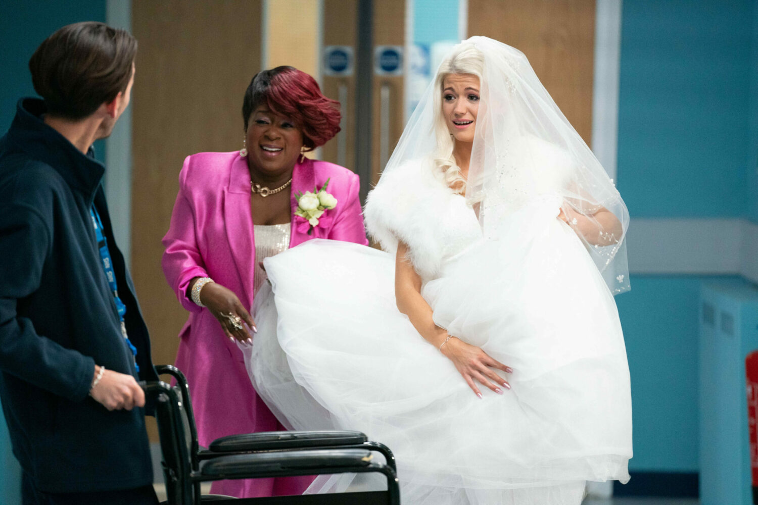 Lola is wearing a wedding dress in the hospital, with Kim helping her