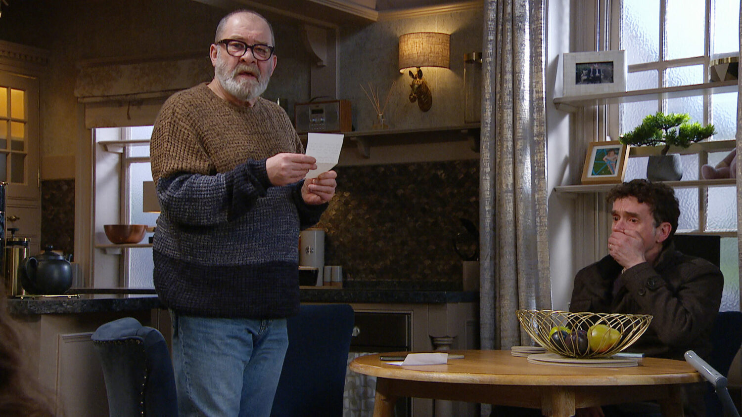 Emmerdale's Bear is holding up a note whilst Marlon is looking upset with his hand over his mouth