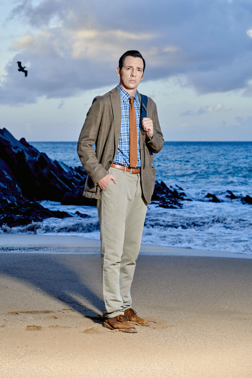 Neville Parker stands on the beach in his suit looking serious in Death In Paradise promo shot
