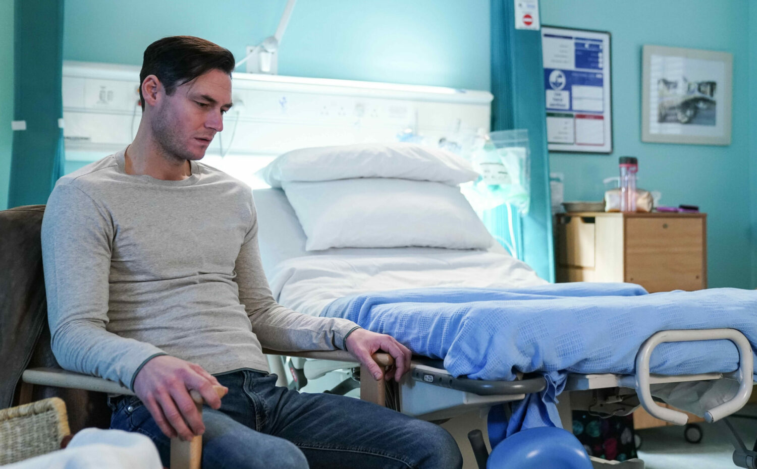 Zack in EastEnders looks upset in a chair next to a hospital bed