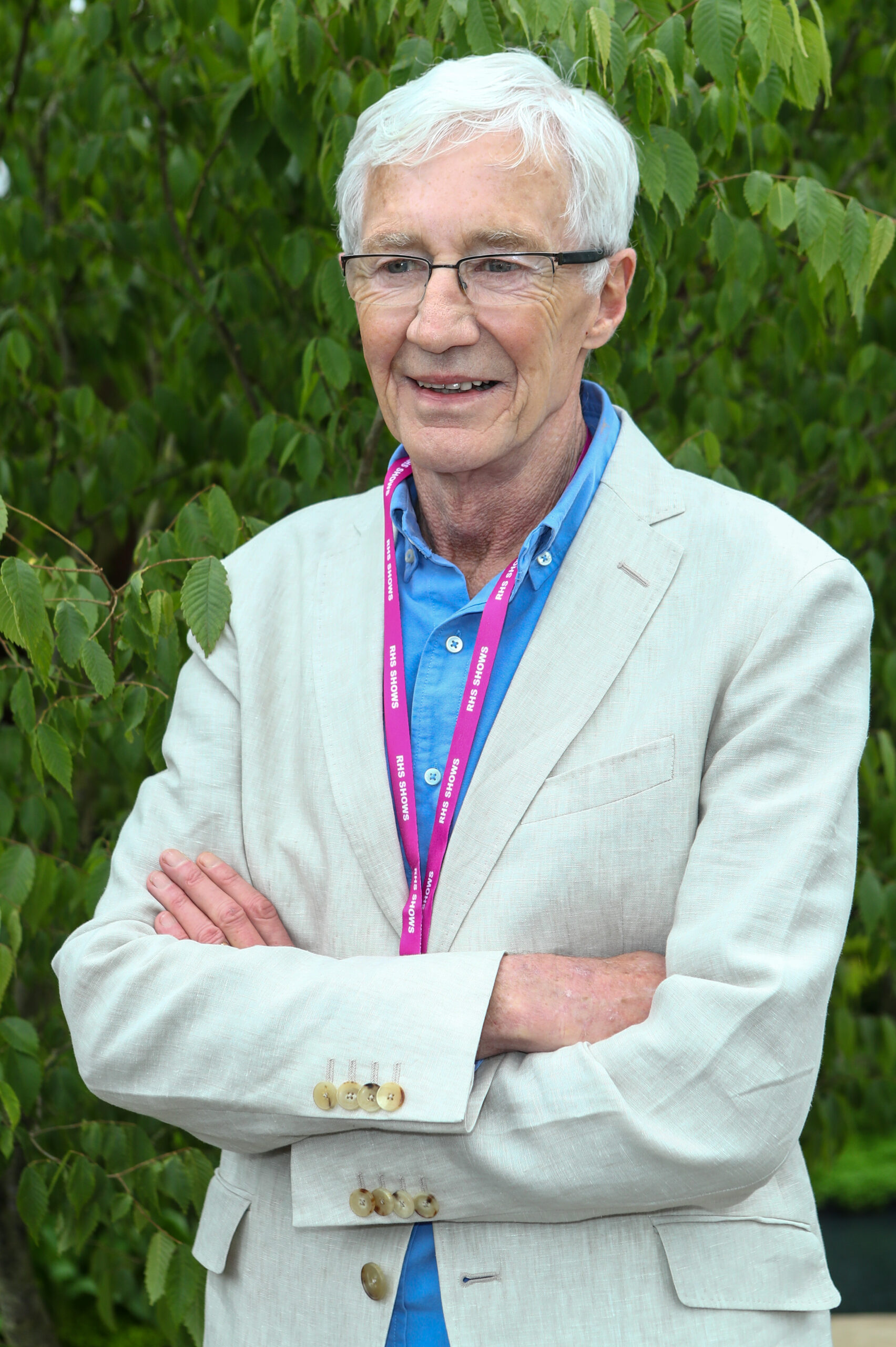 Paul O'Grady smiles with his arms folded in front of a bush at the Chelsea Flower Show