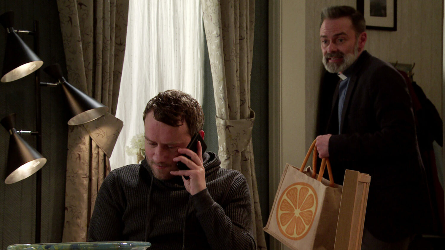 Paul on the phone in Corrie as Billy walks into the room, carrying a bag