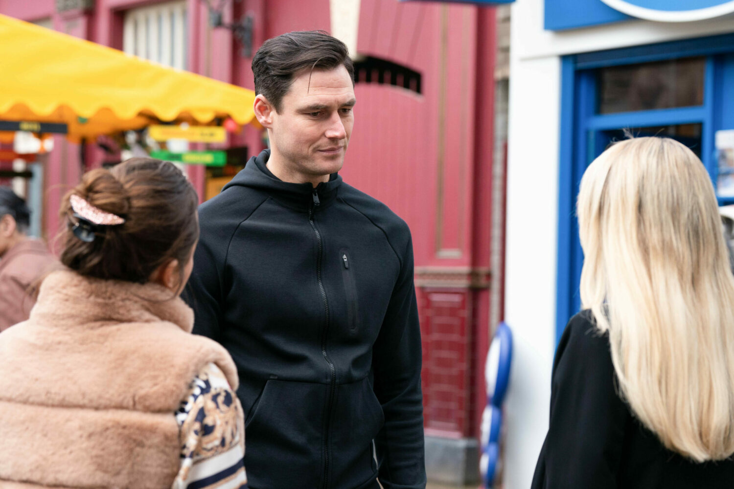 Zack smiles as he talks to Sharon and Whitney in the Square on EastEnders