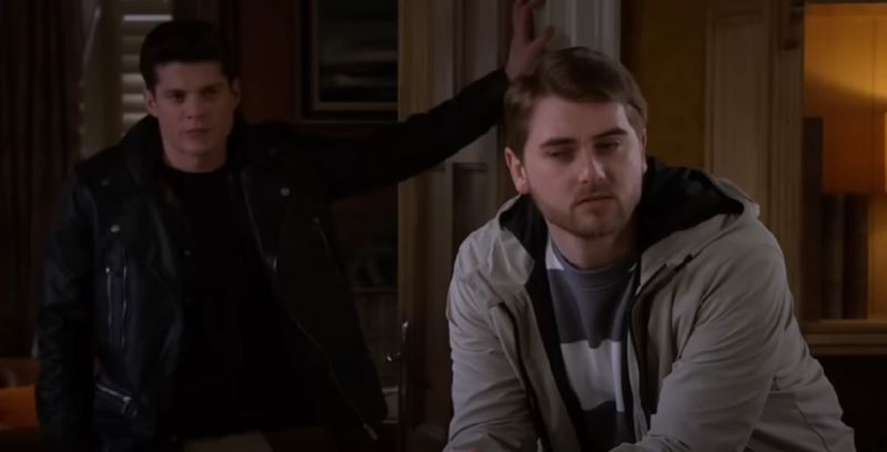 Nicky stands in the background as his mystery visitor talks, looking solemn on Emmerdale