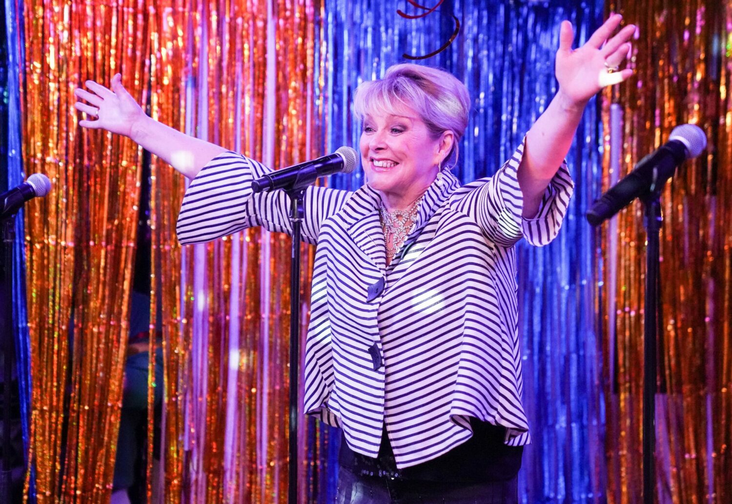 Cheryl Baker throws her arms out wide smiling behind a microphone in EastEnders