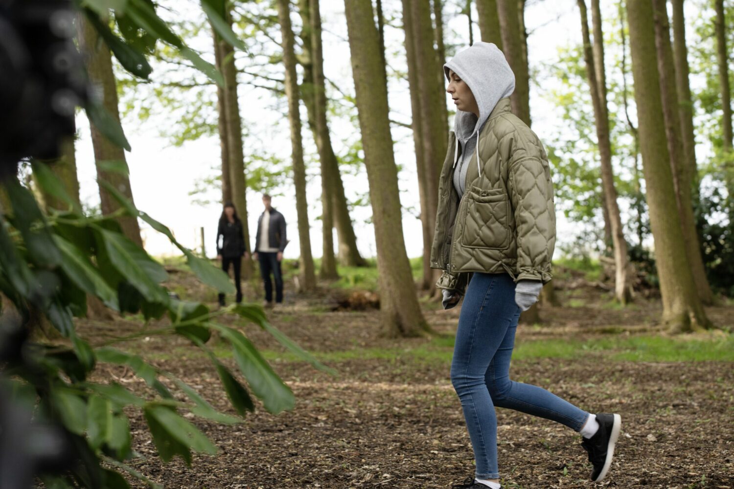Emmerdale's Belle is walking through the forest with her hood up, with Chas and Tom looking on in the distance