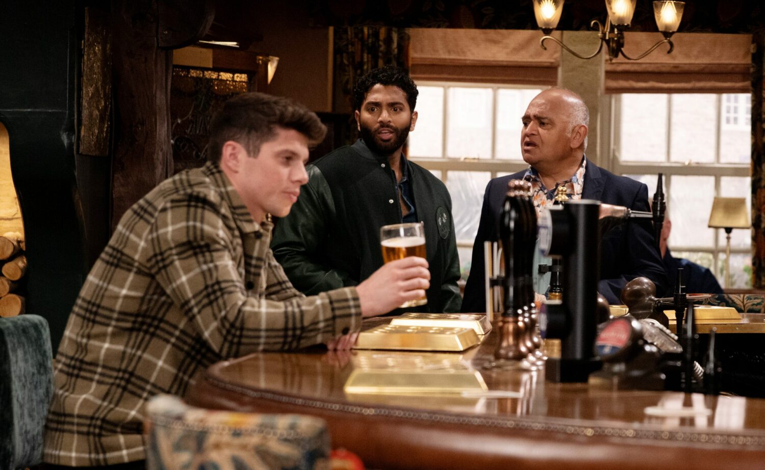 Nicky Miligan sits at the bar of the Woolpack drinking a pint while Suni and Rishi look at him