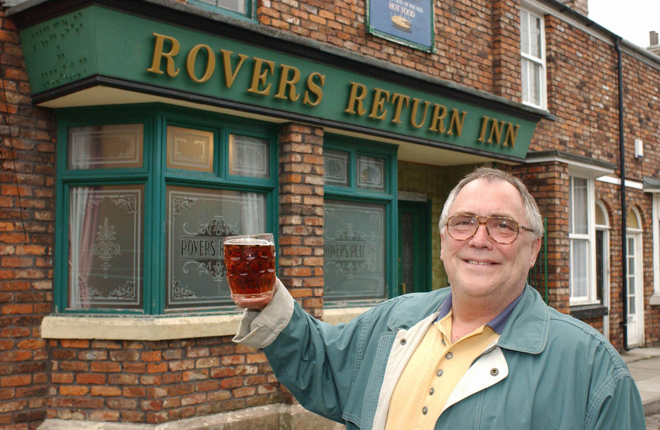 Jack Duckworth standing in front of the Rovers Return in Coronation Street holding up a pint