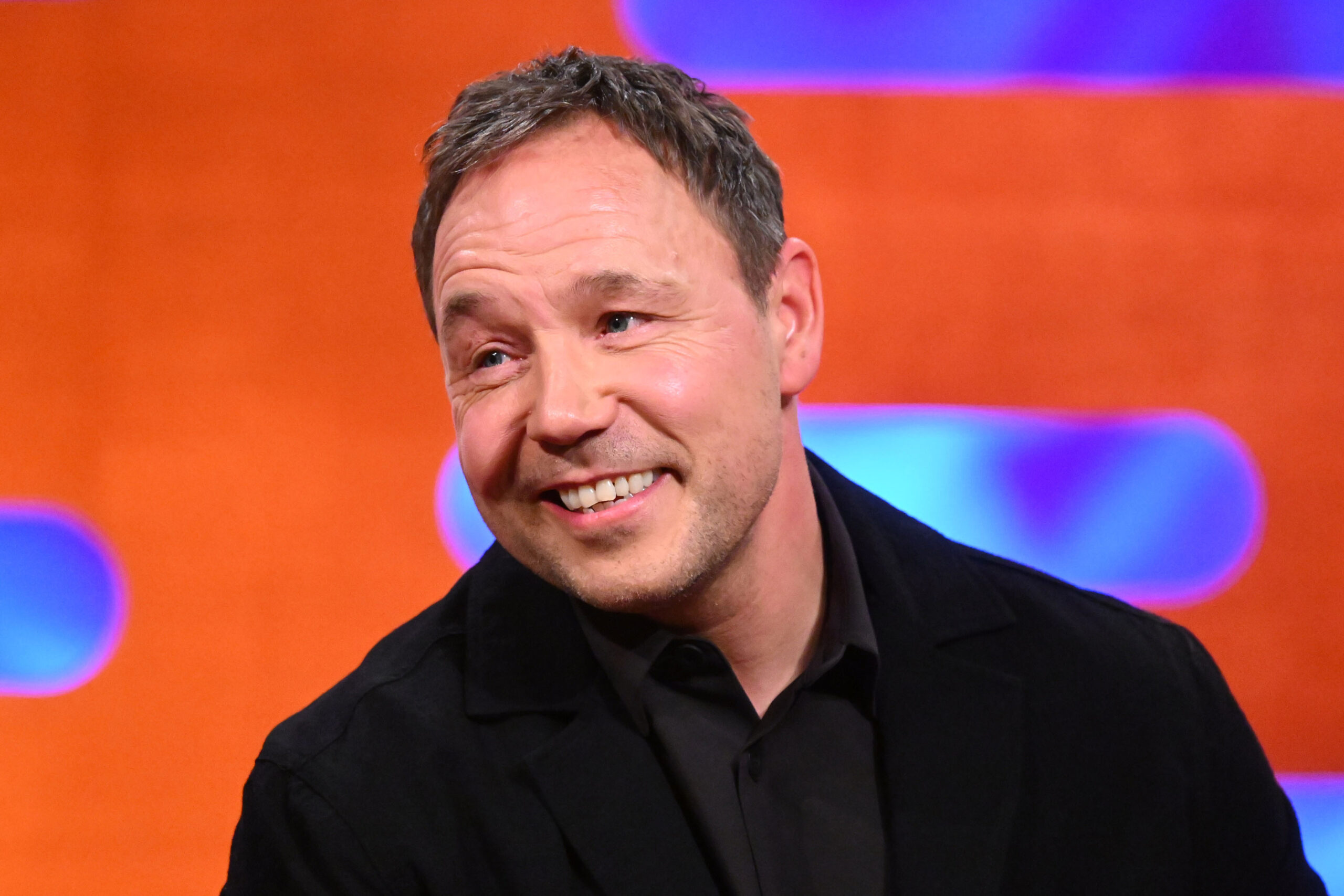 Stephen Graham smiles wearing a black blazer and shirt