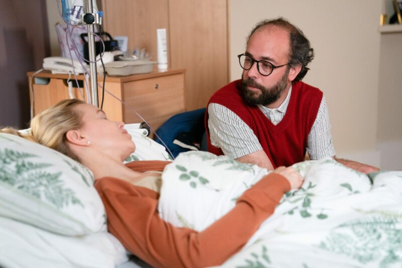 Debbie in hospital while Reiss looks over her on EastEnders