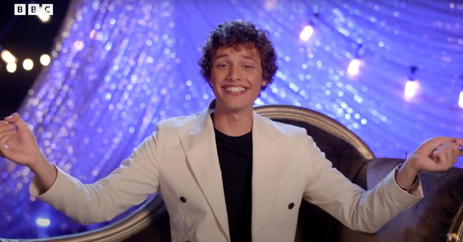 Bobby Brazier speaking in an interview about appearing on Strictly Come Dancing 2023. He is wearing a beige blazer and is sat down in front of a purple backdrop