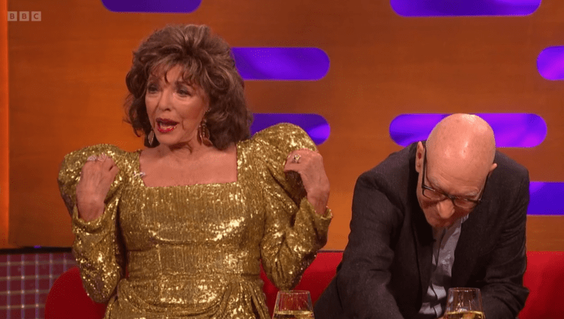 Dame Joan Collins touches her shoulders as Patrick Stewart leans down on the Graham Norton sofa