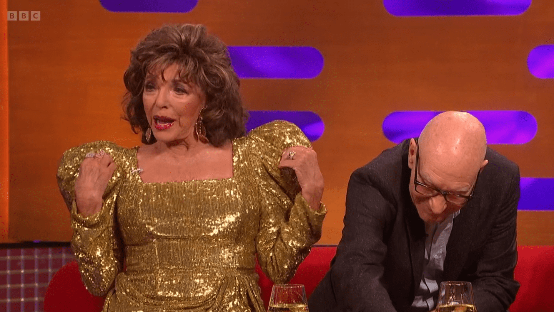 Dame Joan Collins touches her shoulders as Patrick Stewart leans down on the Graham Norton sofa